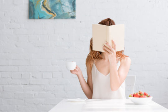 Woman with cup of coffee and bowl of strawberries covering face with book at home